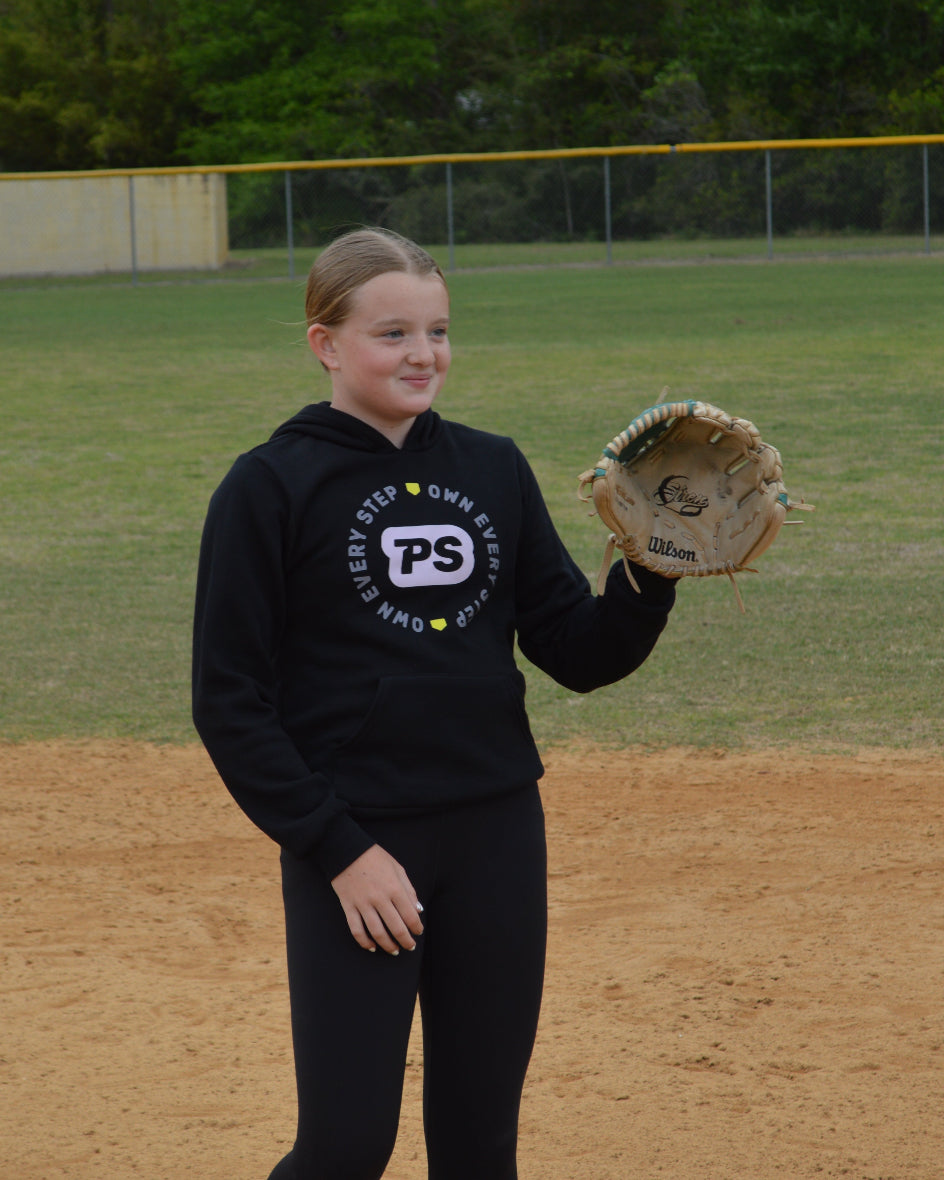 Young girl wearing a black hoodie with a Prep step softball apparel logo design, holding a softball glove on a softba field.