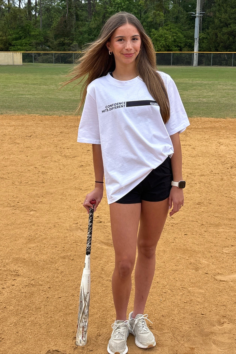 Girl wearing Heavyweight Premium white Tshirt with Confidence Hits Different and Prep Step Softball Apparel logo in a dark purple while leaning against a softball bat and standing on a softball feld